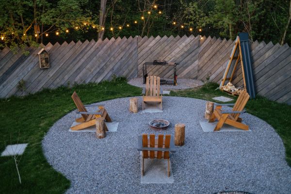 View into a small backyard with gravel fire pit zone, glowing lamp, layered plants, and wood chairs for small backyard ideas without grass.