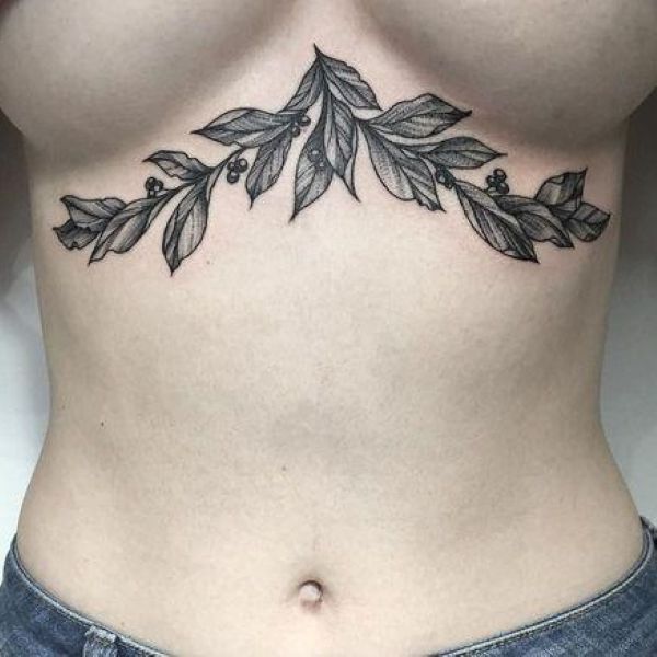Standing mirror shot of sternum and underboob tattoo with ivy vines curving under bust toward ribs, medium wrap design black ink light shading, elegant natural mood.