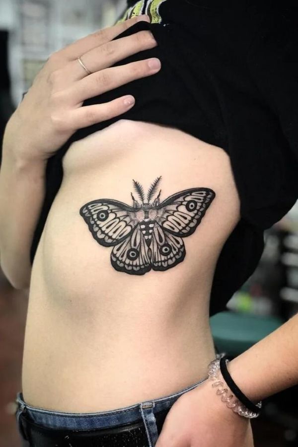 Moth underboob tattoo with bold black wings, symmetrical pattern, and clean linework on rib area.